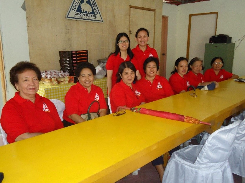 A group of individuals in red shirts with a logo gathers around a yellow table, with eight people seated and two standing behind them. The background features a sign that reads 'Toclong 1st Multi-Purpose Cooperative' and a table with a yellow checkered cloth holding items. This image symbolizes the cooperative's unity and dedication to community growth and development.