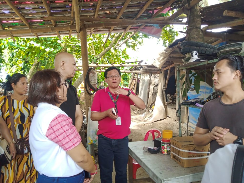 A group of six people are gathered outdoors under a makeshift structure with a bamboo-framed roof. One man, wearing glasses and a red polo shirt, is gesturing with his hands while holding a smartphone. In front of him is a table with jars, boxes labeled "SHOCK ABSORBER," and other small items. The people around him seem attentive.