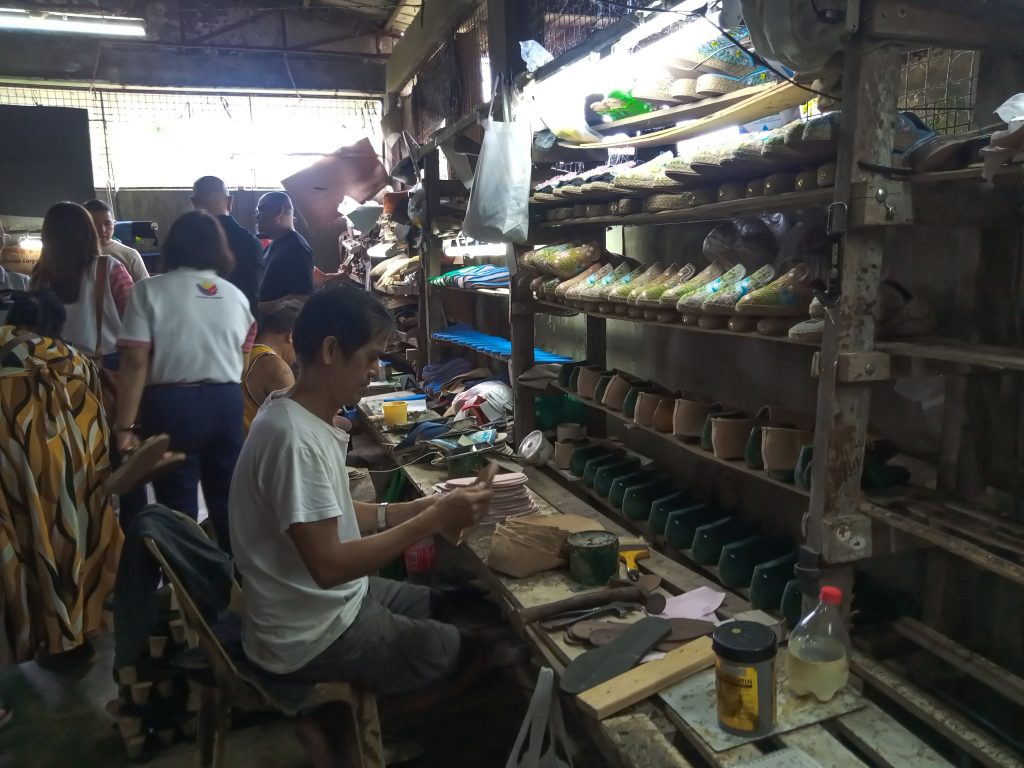 Shoemaker at work in small workshop, surrounded by shoe parts and onlookers.