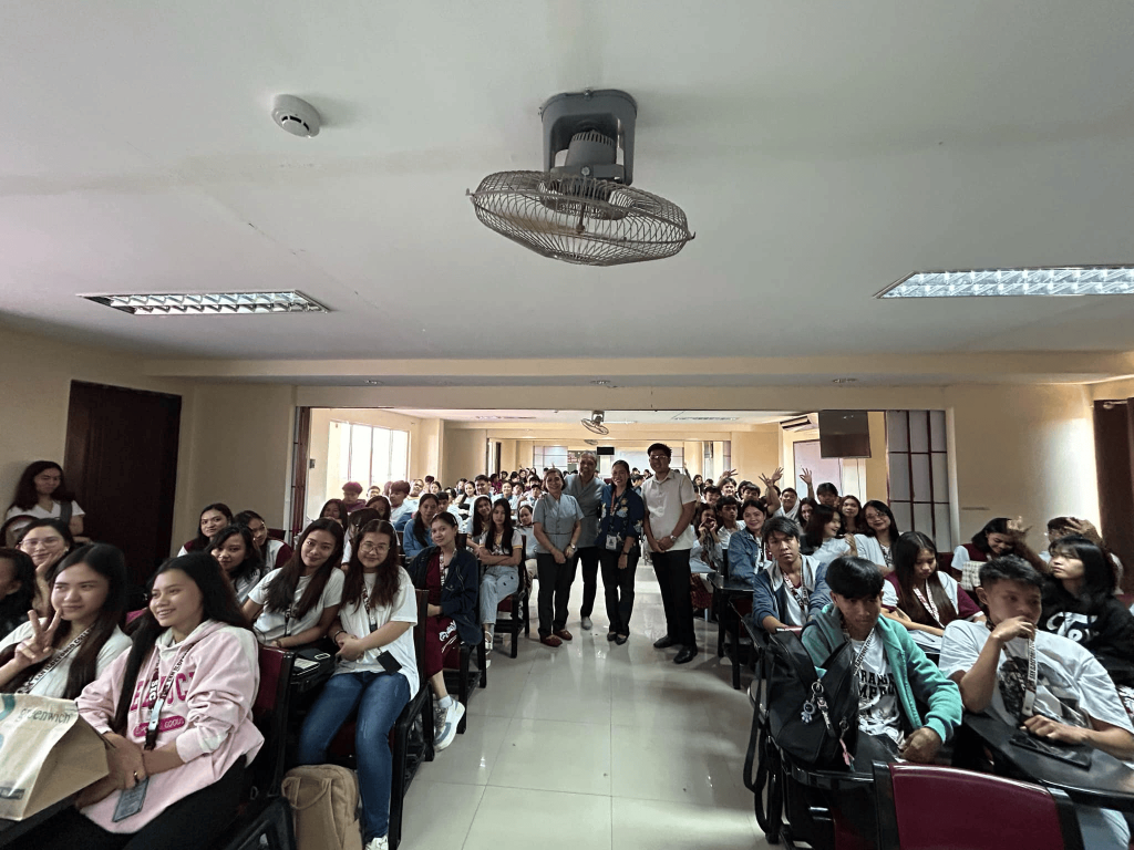 A classroom filled with smiling students seated in rows, with two men standing in the center aisle and a ceiling fan overhead, capturing a lively lecture or workshop atmosphere.