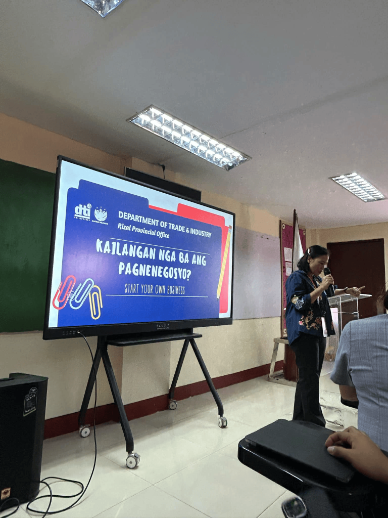 A speaker stands at a podium next to a large screen displaying a DTI Rizal Provincial Office presentation titled “KAILANGAN NGA BA ANG PAGNENEGOSYO? Start your own business.”