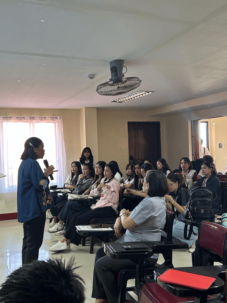 A woman in a blue top stands at the front of a classroom, addressing a group of young women seated in chairs, creating an engaging discussion atmosphere.