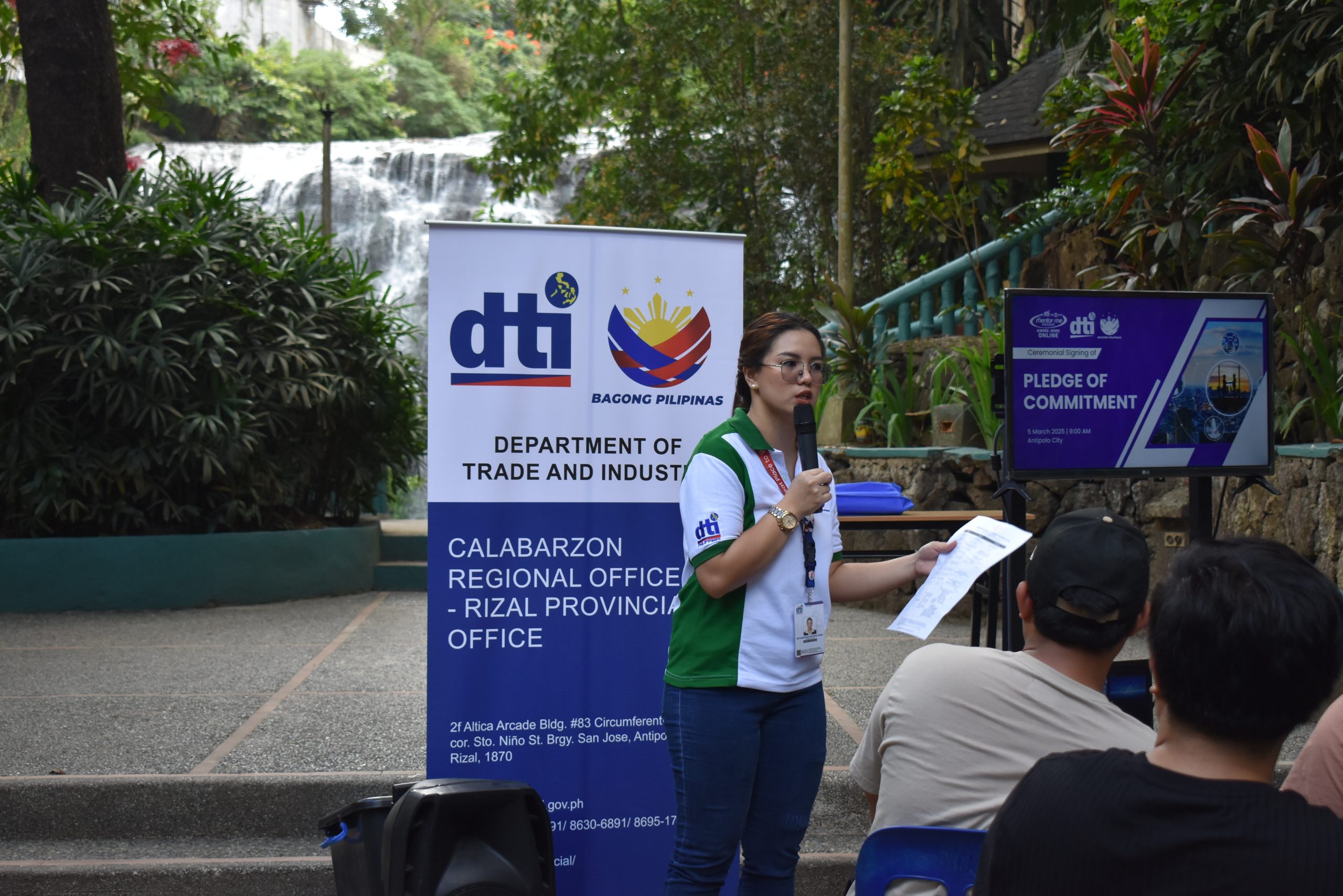 A DTI event taking place outdoors, with a woman addressing a group. The event includes a "PLEDGE OF COMMITMENT" presentation, as shown on the screen, and the DTI CALABARZON banner is visible. The location features a waterfall, suggesting an outdoor or nature-related gathering.