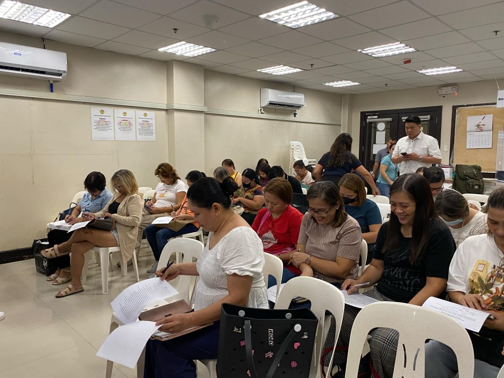 A group of people, who appear to be mostly women, are seated in white plastic chairs in an indoor setting, possibly attending a workshop or meeting. They are writing on papers and appear to be engaged in an activity. The room has fluorescent lighting and several papers pinned to a board on the wall, suggesting an informational or instructional setting. An air conditioning unit is visible on the wall. The individuals in the image seem to be of diverse ages and are dressed casually. Based on the visual context, they might be participating in a training or educational event.