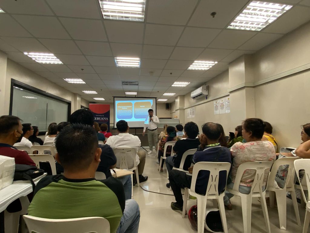 A group of people are seated in white plastic chairs in an indoor setting, possibly a meeting or training room. They are facing a screen where a presentation is being shown, and a person is standing at the front, seemingly giving a presentation. The room appears to be well-lit with fluorescent lighting and has an air conditioning unit on the wall. The individuals in the image seem to be of diverse ages, and based on visual cues, may represent a variety of ethnic backgrounds. They are dressed in casual attire. The setting suggests an informational or instructional event, potentially a seminar or workshop. A banner with the word 'CEBUANA' is partially visible on the left side, which might indicate a connection to a company or organization. The audience appears to be engaged and attentive.