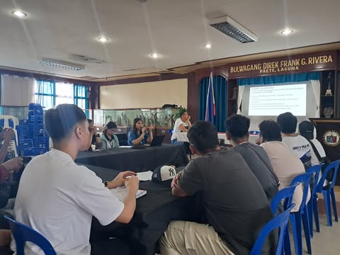 A group of people are seated at tables facing the front of a room, where a presentation is being given. A sign reading 'BULWAGANG DIREK FRANK G. RIVERA PAETE, LAGUNA' is visible. A woman is speaking at the front of the room, near a projector screen displaying text. The attendees are seated in blue plastic chairs.