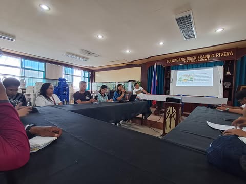 A group of people are seated around a long, dark-colored table facing the front of a room, where a presentation is being given. A projector screen displays text and graphics. A sign reading 'BULWAGANG DIREK FRANK G. RIVERA PAETE, LAGUNA' is visible above the screen. The room has a light-colored ceiling with visible fixtures and blue-framed windows.