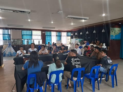 A group of people are seated around several tables covered with black cloth, facing the front of a room. They appear to be attending a meeting or presentation. Blue plastic chairs are visible. The room has a light-colored ceiling with visible fixtures, and blue-framed windows in the background. A world map is visible on a wall in the background.