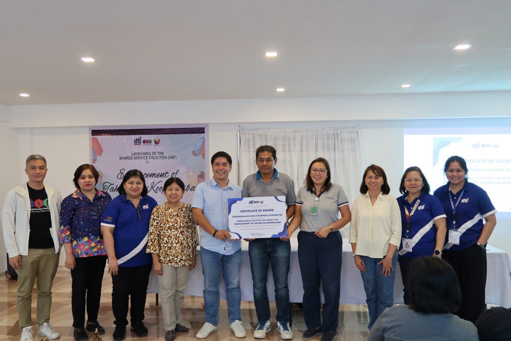 A group of nine people are posing for a photo indoors, holding a framed certificate. They are standing in front of two banners, one of which reads 'Launching of the Shared Service Facilities (SSF)'. The certificate appears to be an award. The people are diverse in age and appearance, and some are wearing matching polo shirts with a logo. A portion of a seated audience is visible in the foreground.