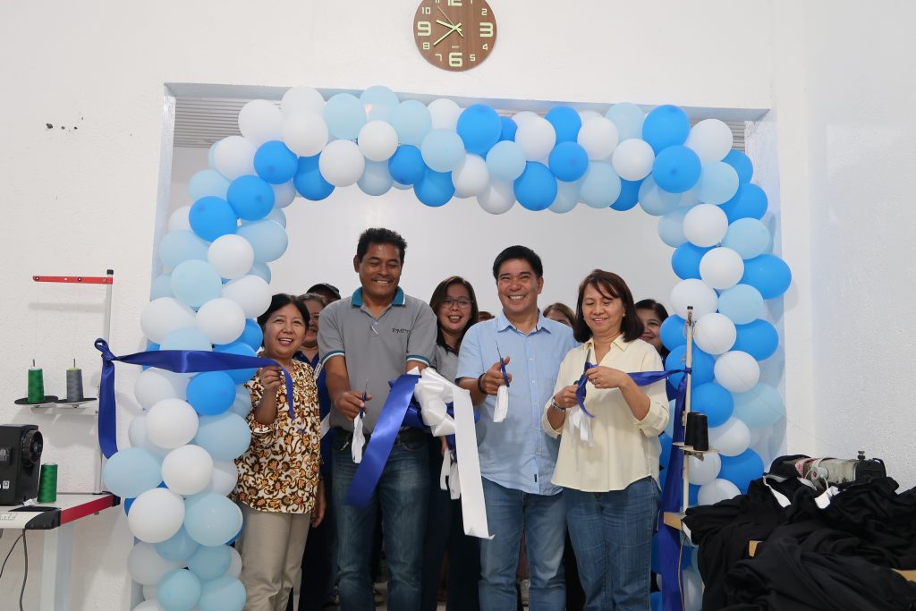 Four people are cutting a blue ribbon stretched across an archway made of blue and white balloons. They appear to be celebrating an opening or launch event. Two men and two women are smiling and holding scissors. A clock is visible on the wall in the background. Sewing machines and fabric are also visible in the room.