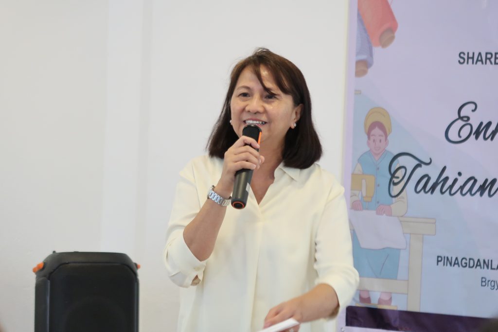 A woman with short brown hair, wearing a light-colored top and a watch, speaks into a microphone. She appears to be giving a presentation or speaking at an event. A black speaker is visible in the lower left corner. A partially visible banner in the background features text that includes 'Enhancement of Tahanan' and 'PINAGDANLA'.