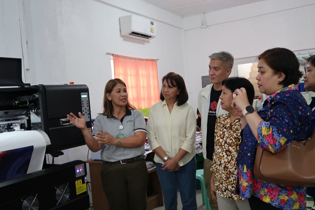 A group of five people are standing indoors, gathered around a large piece of machinery, possibly a printer. One woman, wearing a gray polo shirt, appears to be explaining or demonstrating the machine, gesturing with her hands. The other people are listening attentively. The room has white walls and a window with orange curtains. An air conditioning unit is visible on the wall.