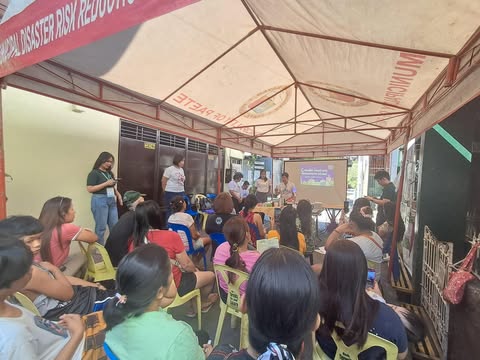 A community event or workshop, possibly related to "Disaster Risk Reduction" (as suggested by the sign), is being held outdoors under a tent. Presenters are using a screen to display information to the attendees.