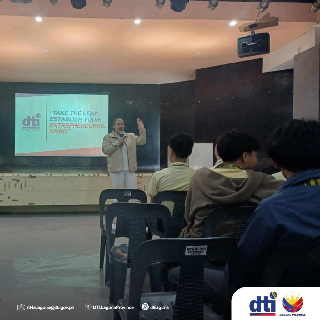 A resource speaker stands on stage delivering a presentation titled "Take the Leap: Establish Your Entrepreneurial Spirit" (visible on the screen) to seated participants during the DTI-Laguna Seminar on Budget Management in Santa Rosa City.