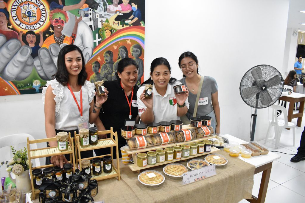 Four women are standing behind a table displaying various food products in jars and containers. A colorful painting is visible in the background. The text "Rizal" and some names are also visible in the image.