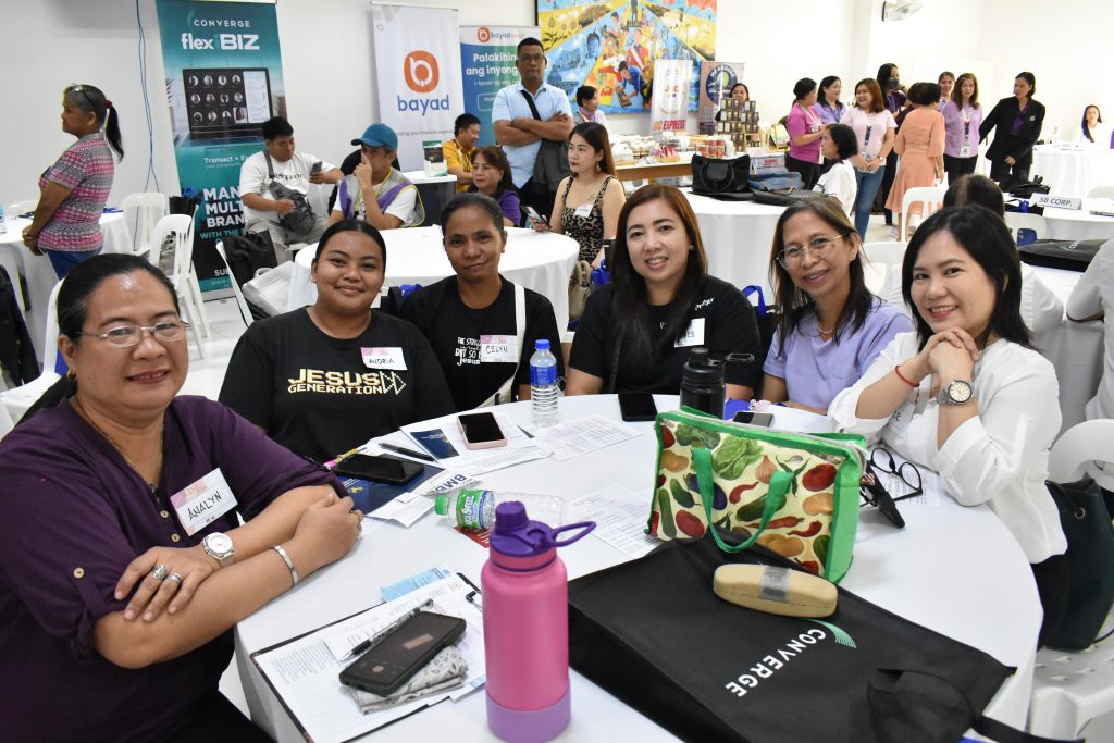 Six women are sitting around a table with Converge materials, and a display of products is visible in the background. The image also includes text elements such as "CONVERGE," "flex BIZ," "Palokihin ang inyong bayad," and "MAN MULTI BRANCH."