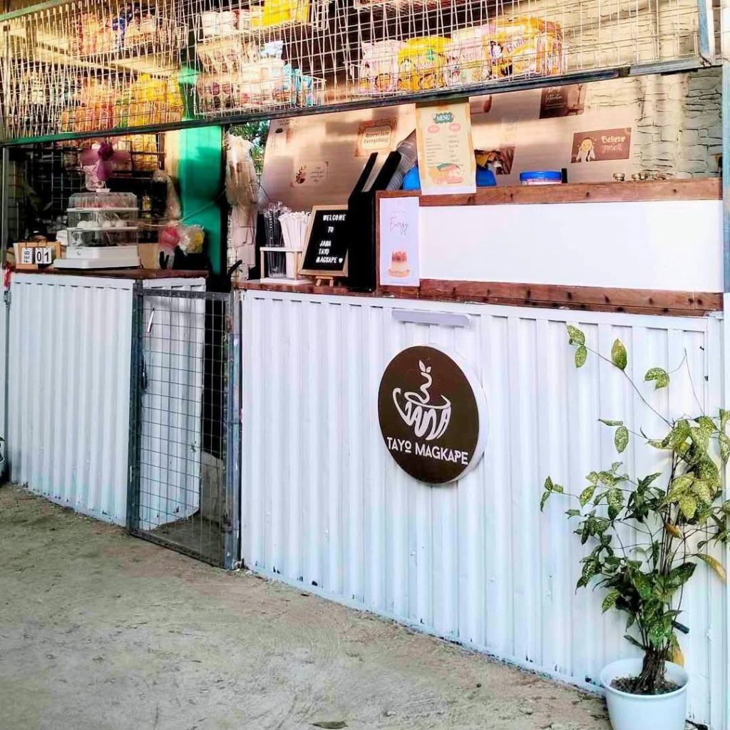 Small coffee shop named "Tayo Magkape" with a white corrugated metal counter, wooden top, steaming cup logo, and various snacks and drinks displayed behind the counter; a potted plant sits to the right
