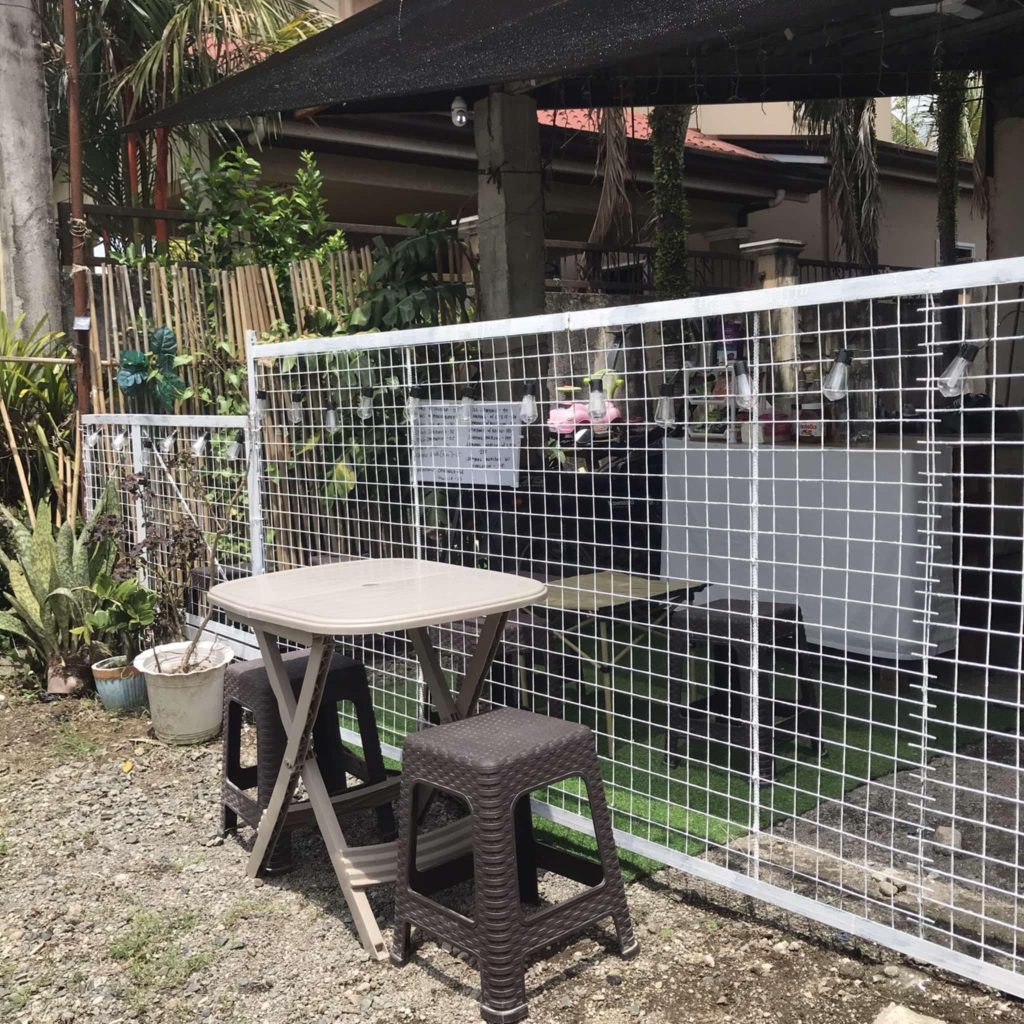 Outdoor seating area with a small table and two brown stools in front of a white wire fence, surrounded by potted plants and hanging decorations, with a house and shaded garden in the background