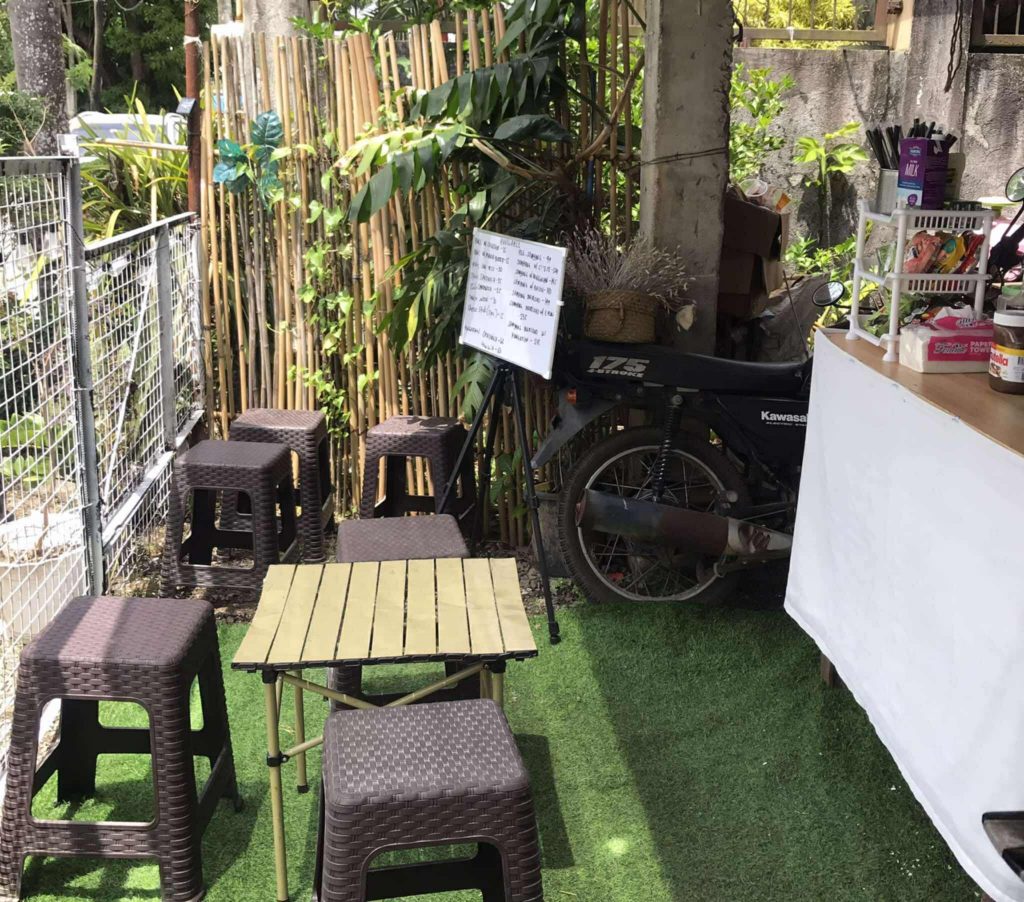 Outdoor seating area with a small wooden table and six brown plastic stools on artificial grass, next to a bamboo fence and green plants; a white handwritten menu sign listing coffee options is propped against a black motorcycle, with a counter displaying utensils and condiments nearby