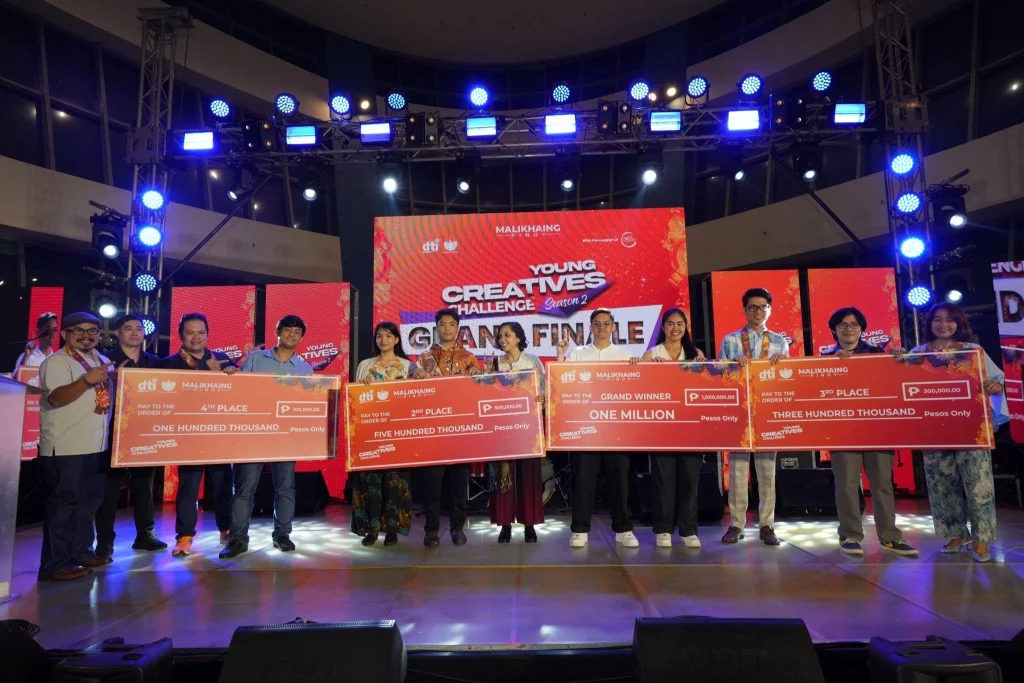 Young Creatives Challenge Grand Finale" event. The stage is brightly lit with multiple spotlights and a large red banner in the background displaying the event name. The checks have various amounts written on them, including "One Million," "Five Hundred Thousand," and "One Hundred Thousand.