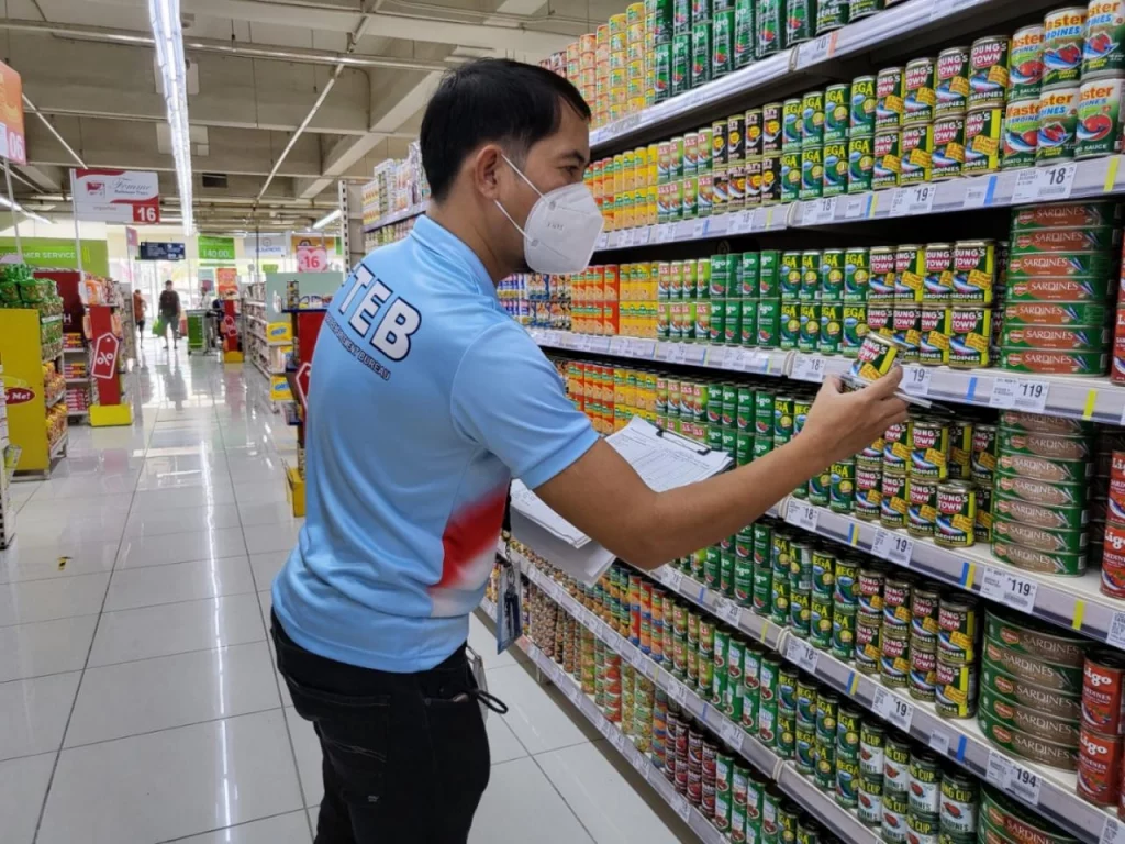 A DTI field officer wearing a blue  shirt checks canned goods in a grocery store aisle using a checklist, as part of the agency’s nationwide price freeze enforcement and supply monitoring in calamity-affected areas.
