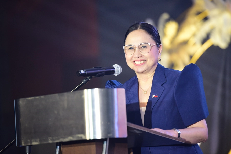 DTI Secretary Cristina A. Roque stands behind a podium, speaking into a microphone. She is wearing a blue outfit with puffed sleeves and a Philippine flag pin on her chest. The background is dark with blurred golden decorations, reflecting a formal setting during the announcement of PHP 66 billion in ecozone investments in Pasay City, Philippines.