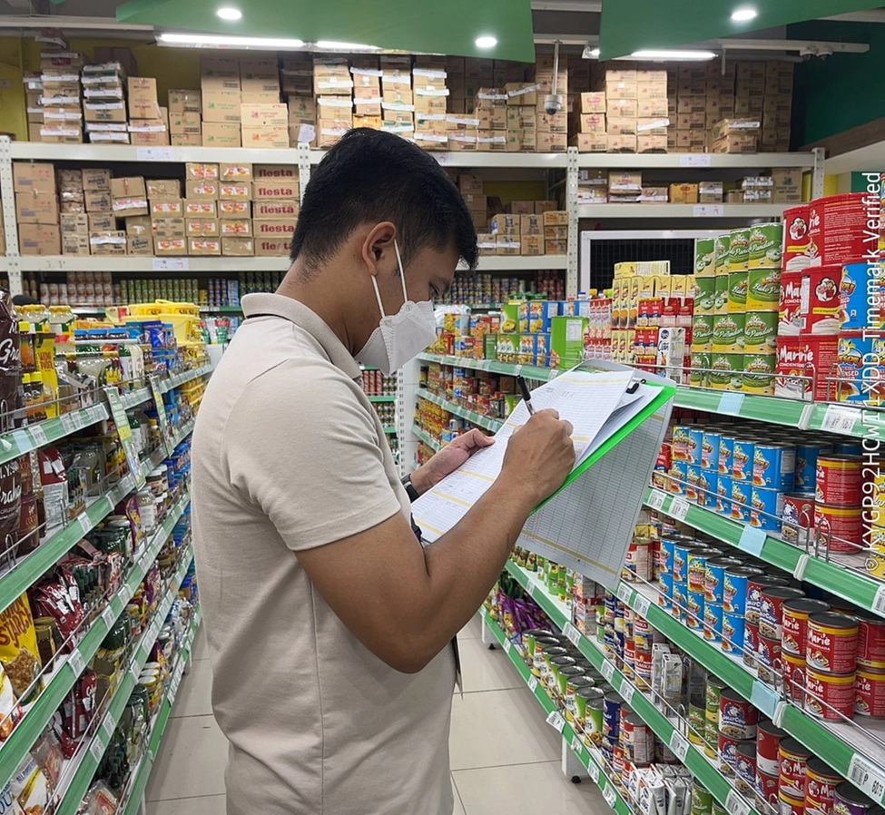DTI personnel conducting price and supply monitoring in a grocery store aisle in Eastern Visayas, following the state of calamity declaration. The individual is wearing a white polo shirt and face mask, holding a clipboard while inspecting shelves stocked with canned goods and other essentials.