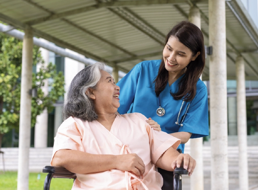 A healthcare professional in blue scrubs converses with an older patient in a wheelchair under a covered outdoor walkway.