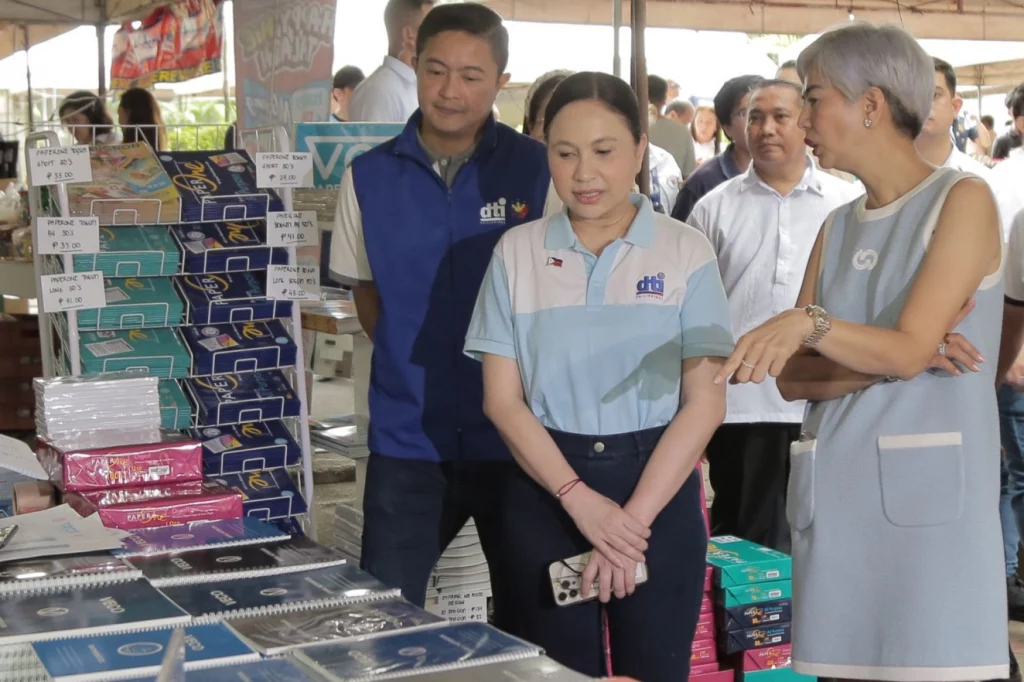 DTI officials and shoppers at the Diskwento Caravan: Balik-Eskwela Edition in Manila, browsing discounted school supplies displayed on tables under a tented area.