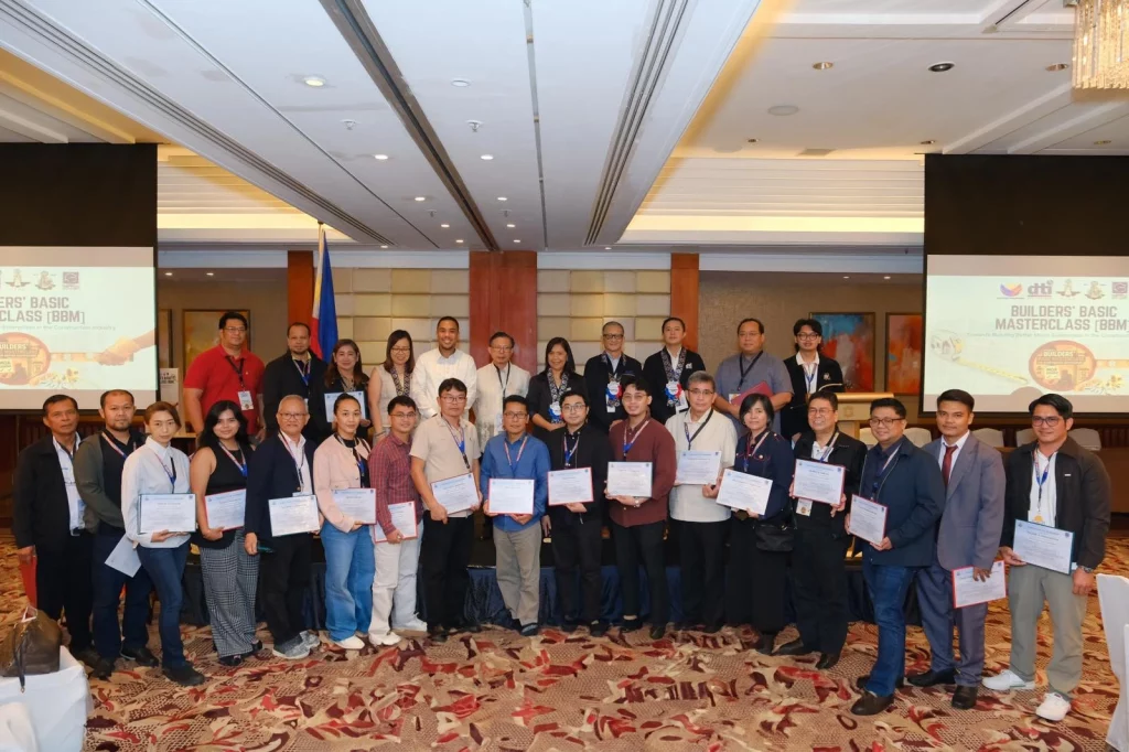 Group photo of participants at the DTI's Builders’ Basic Masterclass (BBM) for pakyaw contractors. Around twenty individuals pose in a conference room holding certificates, with two large screens in the background displaying 'Builders Basic Masterclass (BBM)' and the DTI logo. The setting reflects a formal training event.