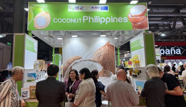A booth at TuttoFood Milano showcasing coconut-based products from the Philippines. The booth features a large banner labeled 'COCONUT Philippines' with coconut imagery. Several attendees engage with exhibitors and examine various coconut products displayed on shelves, with other international booths visible in the background.