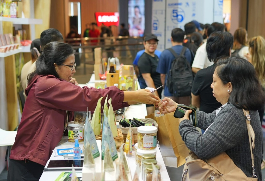 A vendor at the DTI-MisOr ONE Expo 2025 in Makati hands a product to a customer at a busy market stall showcasing MSME goods, with various jars, packets, and bottles on display. Several attendees queue behind the customer, highlighting strong public interest and engagement.