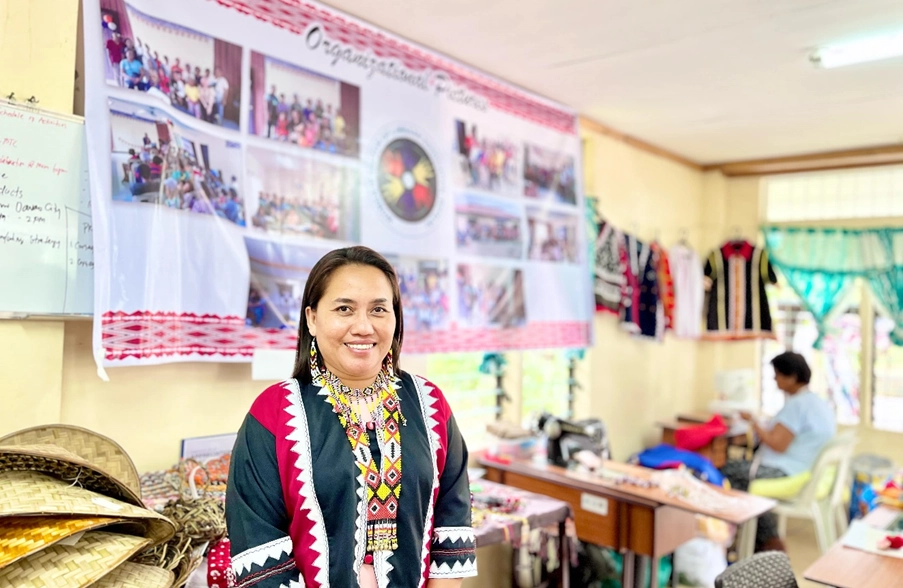 A smiling woman from an indigenous community wears a brightly colored, intricately embroidered garment in a workshop. Behind her, a large banner displays photos of community activities, and various woven crafts and traditional attire are visible, reflecting the group’s dedication to preserving local culture and craftsmanship. A second person is seen working on textiles in the background.