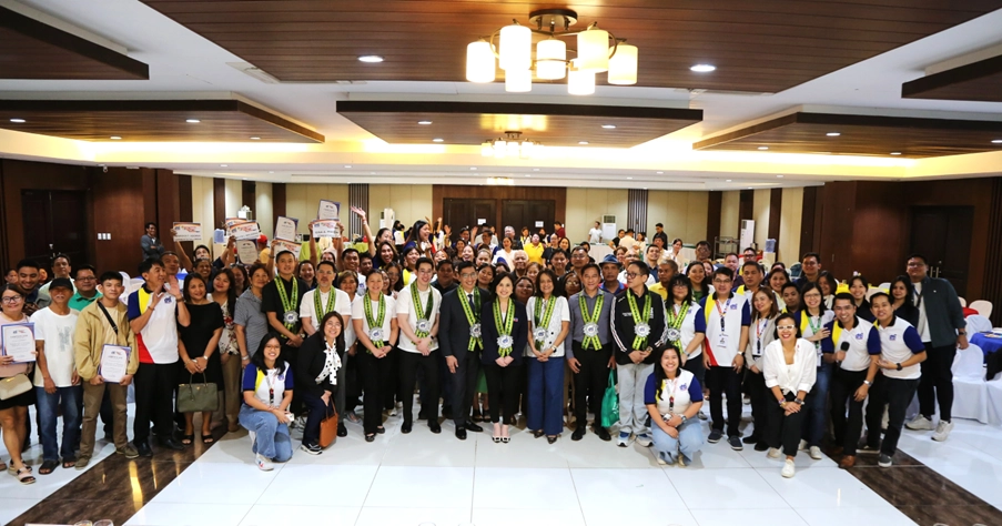 A large group of individuals, who appear to be mostly of Southeast Asian descent based on visual cues, are posing for a group photograph indoors. They are gathered in a spacious room with warm lighting, likely a ballroom or large meeting hall, given the wooden paneling and chandeliers. Many individuals in the foreground are holding certificates or waving small signs, suggesting an awards ceremony or recognition event. Some individuals in the front rows are wearing lanyards with what appear to be identification cards. The overall atmosphere seems celebratory and positive. Based on the title and body of the article, this gathering likely includes Department of Trade and Industry (DTI) Secretary Cristina A. Roque, who may be among those in the front, presenting assistance awards to micro, small, and medium enterprises (MSMEs) in Central Luzon, Philippines. The large number of attendees suggests a significant regional event focused on supporting local businesses.