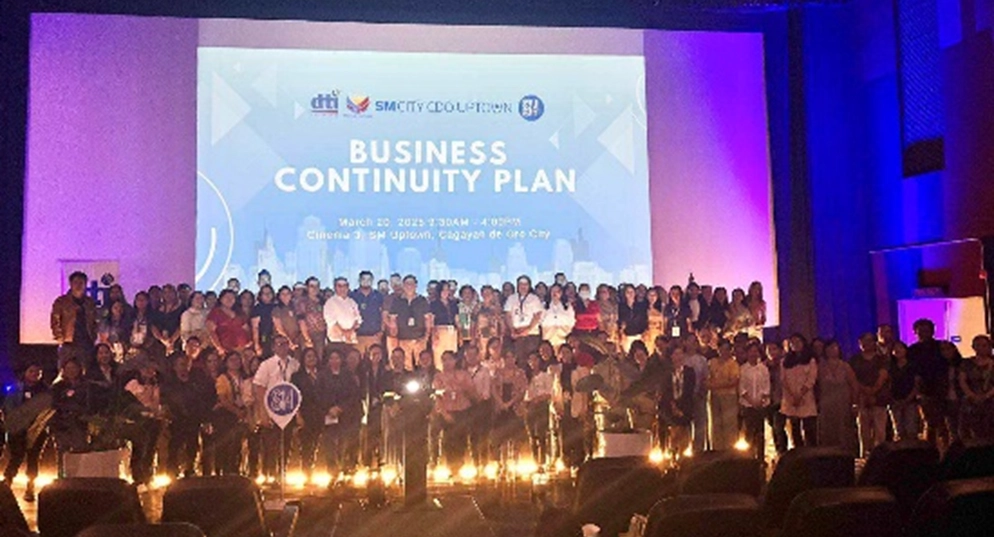 Group photo of participants (MSMEs and SM tenants) posing on stage after the DTI and SM Prime Business Continuity Plan training session held at SM City CDO Uptown's Cinema 3 on March 20, 2025. The event title screen with DTI and SM logos is visible in the background.