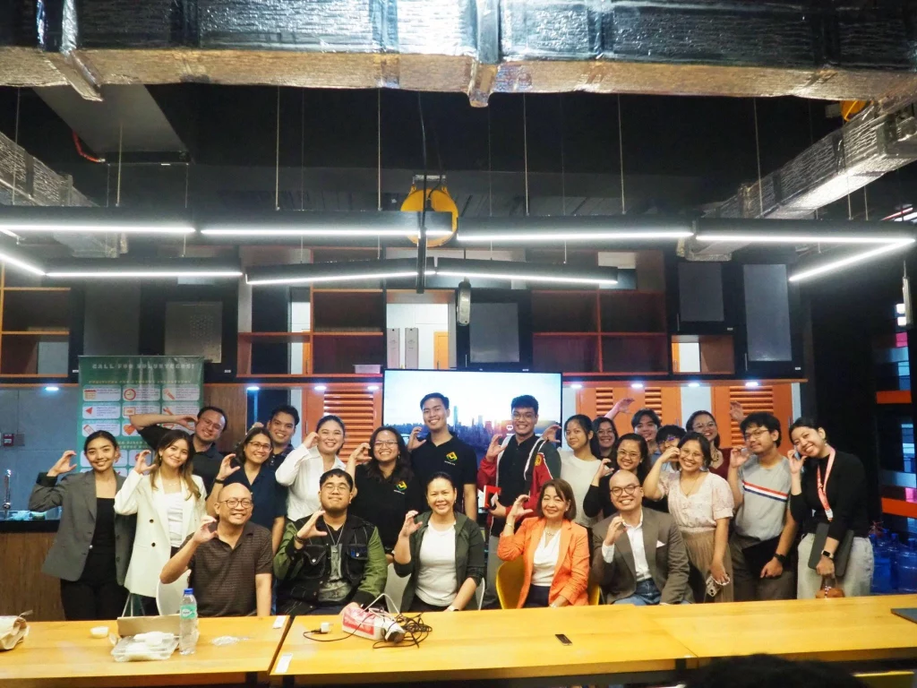 Participants and organizers of the "Malikhaing Pinoy Chats" event pose for a group photo inside the modern, well-lit space of the Benilde Hub of Innovation for Inclusion (HiFi), with many making finger heart gestures.