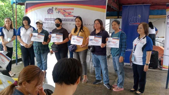 RD Edna Dizon with a group of women stands in a row, each holding a certificate of membership from the DTI (Department of Trade and Industry). They're posing for a picture in front of a banner bearing the DTI logo and the words "Pangkabuhayan sa Pagbangon ng Ginhawa." 