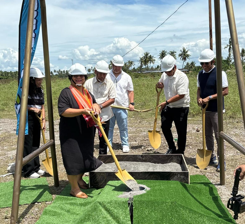 DTI Region 12 Regional Director Flora Gabunales and Coco Island representatives at the groundbreaking ceremony, holding shovels with a backdrop of coconuts, grass, and a scenic sky.