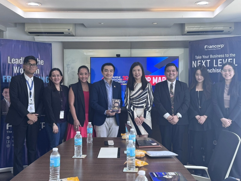Eight individuals are posed behind a conference table in a meeting room. Banners for "Francorp" are visible in the background. The individuals, identified in the caption, include representatives from DTI-FTSC, JIGS, Bench, the Philippine Franchise Association (PFA), and POPMART. One of the POPMART representatives, a man in the center, holds a small, boxy item. Water bottles and papers are on the table. The setting suggests a formal meeting or discussion, likely related to business or trade. Given the context of the article about POPMART's expansion in the Philippines and the presence of DTI and PFA representatives, the people in the image are likely a mix of Filipino and possibly Chinese individuals.