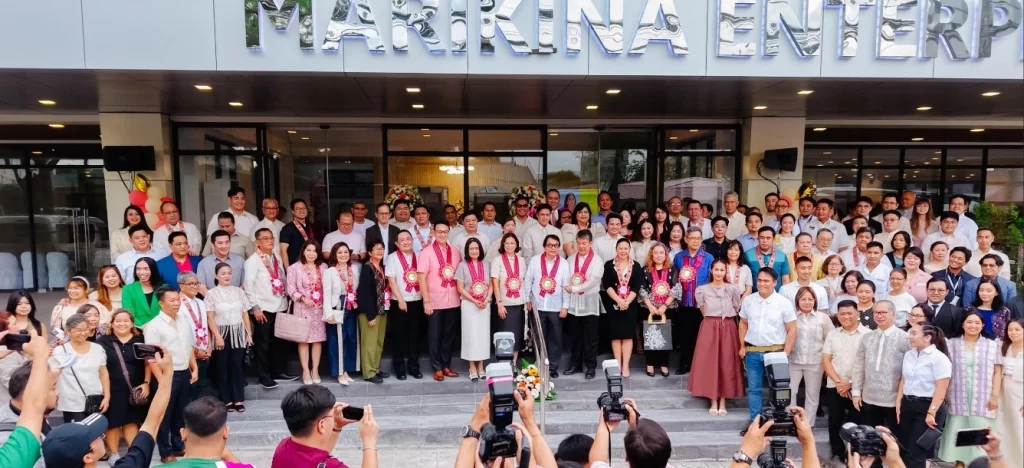 A large group of people, likely over 50, are gathered on the steps in front of a building with "MARIKINA ENTERP" visible at the top. Many individuals in the foreground are holding up phones and cameras, seemingly taking photos of the group. The people in the main group are dressed in a mix of formal and business casual attire. Several individuals in the front row appear to be wearing leis or sashes. The overall atmosphere suggests a formal event or gathering.
