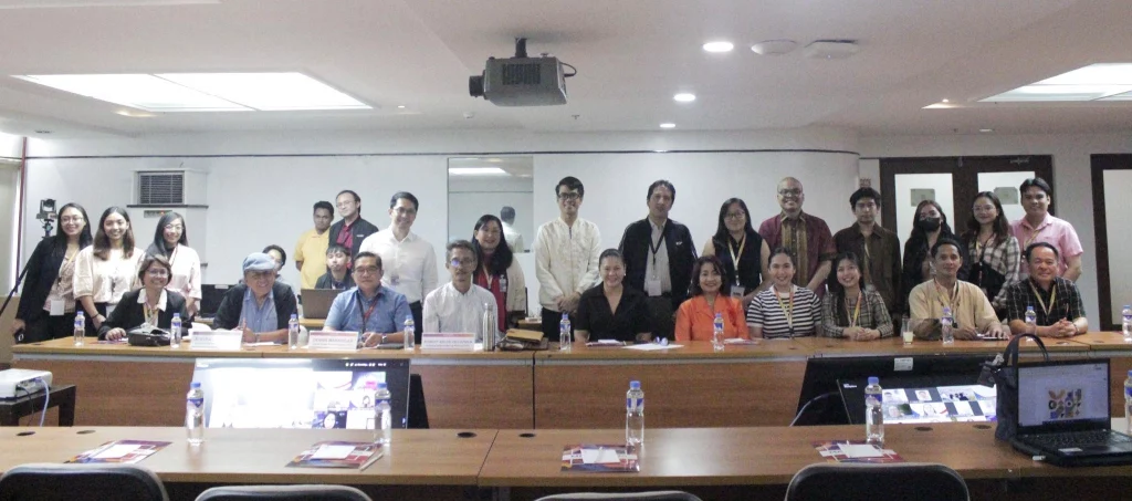 A group of individuals are gathered around a long conference table in a well-lit room. Some are seated and some are standing behind the table. There are laptops, water bottles, and papers on the table. The photo caption identifies some of the people in the image as participants of the “Setting the Stage: Validation Session for the Performing Arts Roadmap,” including individuals from the Cultural Center of the Philippines, Dulaang Unibersidad ng Pilipinas, and the Department of Trade and Industry (DTI).