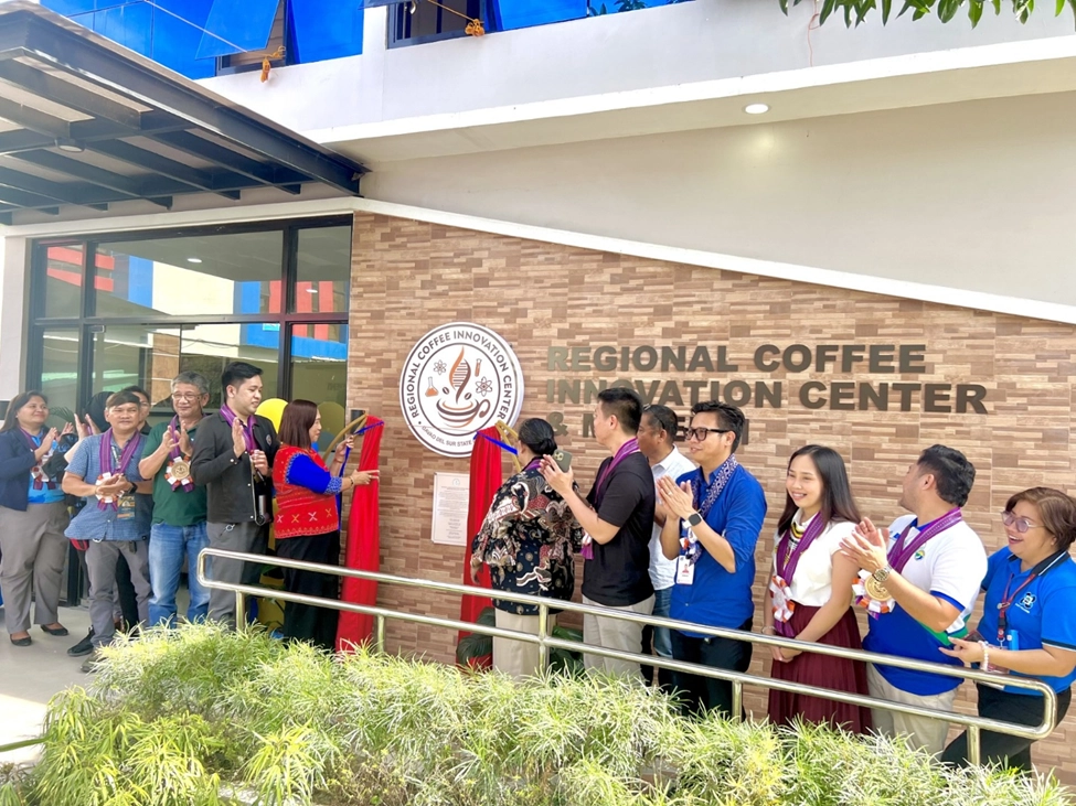 Davao del Sur State College President, Dr. Augie E. Fuentes, leads an unveiling ceremony at the Regional Coffee Innovation Center. He is joined by partners from national government agencies (NGAs), local government units (LGUs), industry, and non-government organizations (NGOs). The group stands in front of the center, with some individuals holding red cloths and others clapping.