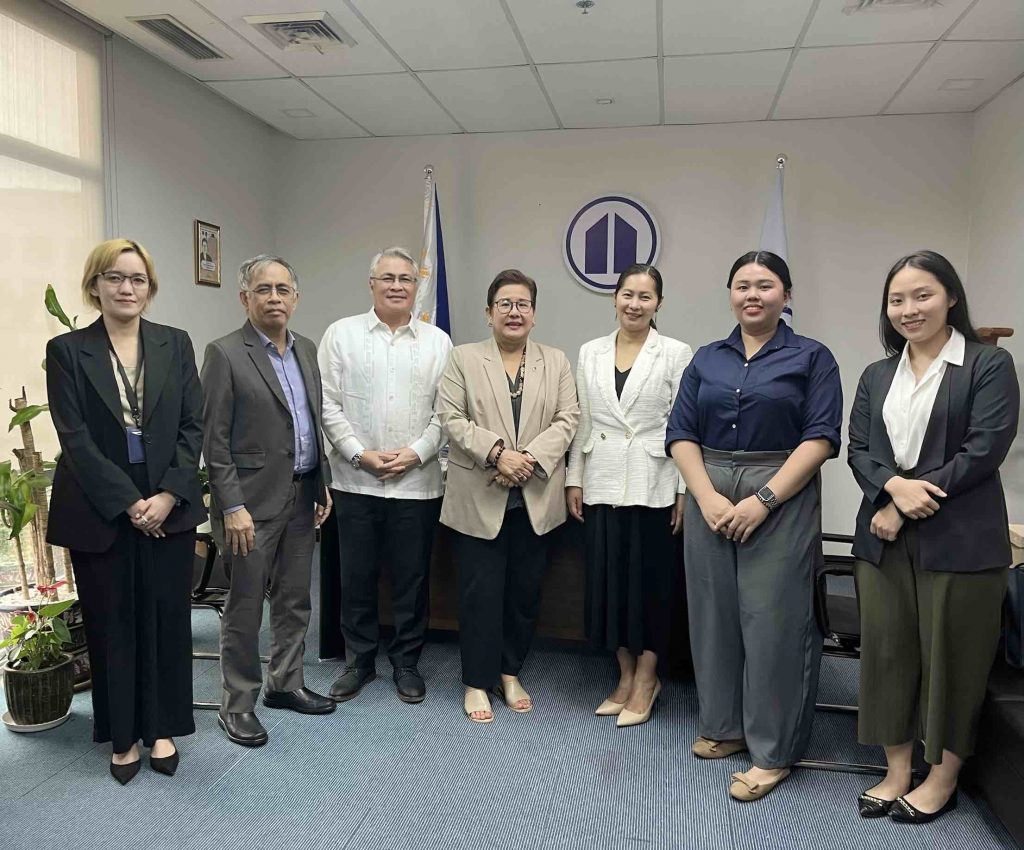 A group photo of seven individuals standing in an office setting, from left to right: PCCI-International Affairs Department Specialist Ms. Francesca Alvarez, PCCI Assistant Secretary General for International Affairs and Chief-of-Staff Mr. Edwin Glindro, PCCI Vice President for International Affairs Mr. Jude Aguilar, PCCI President Consul Eunina Mangio, DTI-FTSC Assistant Secretary Alma Argayoso, FTSC Regional Coordinator Micha Reoyan, and PCCI-International Affairs Department Specialist Chloe Tan. They are standing in front of flags and the PCCI logo.