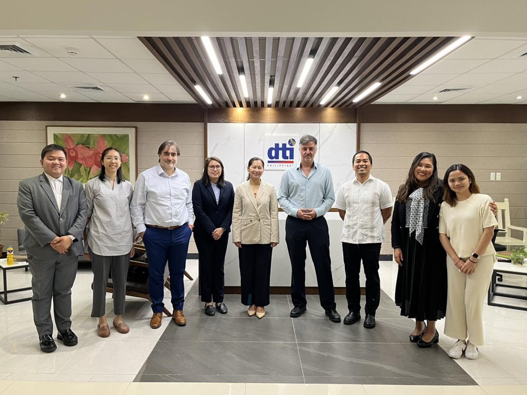 A group of nine individuals stands in a well-lit lobby area in front of a wall displaying the DTI logo. They wear business attire and are posed side by side, smiling at the camera.