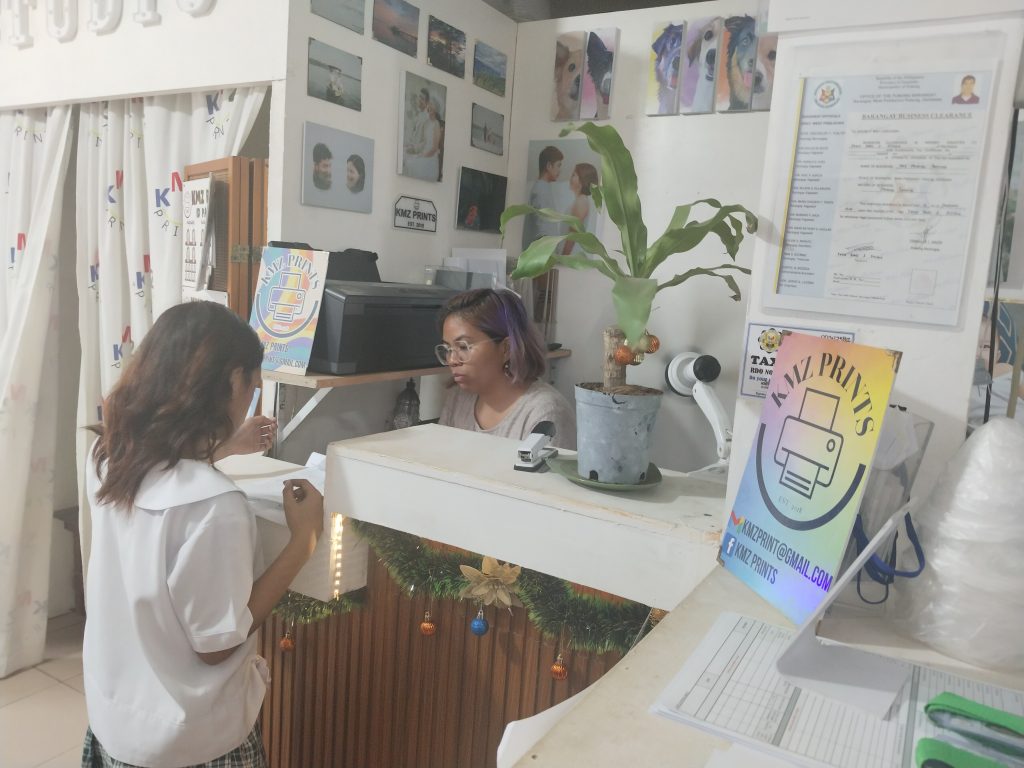 Interior of a photo printing studio with a “STUDIO” sign overhead. A woman with purple-tinted hair is behind the counter working on a computer, while a customer stands in front of the desk. Portrait samples and decorations are displayed on the walls and counter, and a potted plant sits on the reception area.