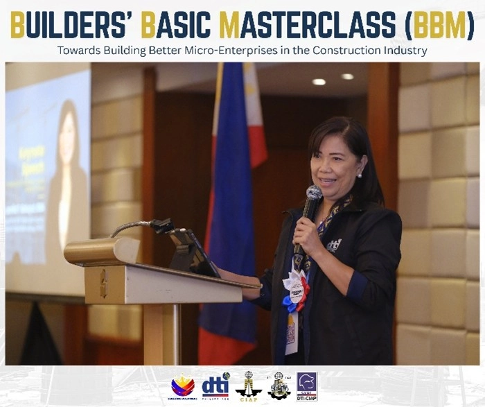 A speaker presenting at the Builders' Basic Masterclass (BBM) launch event, themed 'Towards Building Better Micro-Enterprises in the Construction Industry.' The speaker stands at a podium with a microphone, with a projection screen, the Philippine flag, and logos of partner organizations including DTI, PCAB, and CIAP visible in the background.
