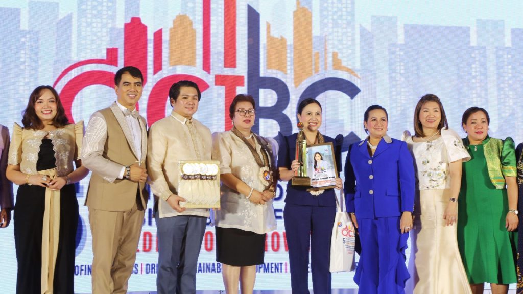 A group of eight people, Ms. Anne Francisco, Dr. Carl Balita, Atty. Arnel Mateo, Consul Enunina V. Mangio, DTI Secretary Cristina A. Roque, Ms. Sarah Deloraya Mateo, Ms. Josephine See and Dr. Gaw So, pose smiling on a brightly lit stage. Some hold awards, with the QCIBC logo visible in the background.