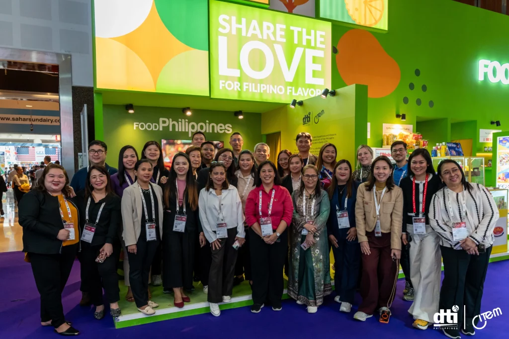 A group of individuals, who appear to be mostly women, are posing for a group photograph in front of a brightly colored booth at an indoor event, possibly a trade show. The booth has the text 'FOODPhilippines' and 'SHARE THE LOVE FOR FILIPINO FLAVORS'. The caption identifies two individuals in the front row as Atty. Anna Grace Marpuri and Chol dela Paz, and mentions they coordinated with exhibitors throughout the trade show. The individuals in the photograph appear to be of diverse ages and possibly of East or Southeast Asian descent, based on visual cues. They are dressed in business casual or professional attire, suggesting they are participants or organizers of the event. The booth and the presence of a 'dti' logo suggest a connection to the Philippine Department of Trade and Industry. The overall setting appears to be a busy and commercial environment, possibly a food-related trade exhibition.
