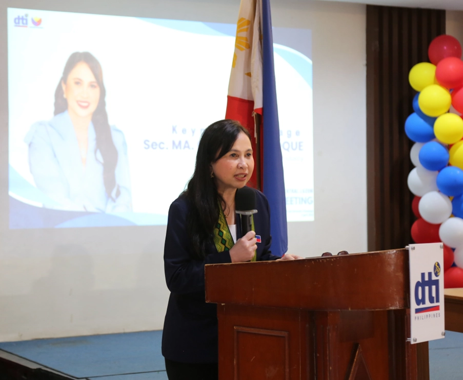 DTI Secretary Cristina A. Roque,  stands at a wooden podium and speaks into a microphone. She is giving a presentation or addressing an audience. To her left, a screen displays a picture of Secretary Roque and text that includes 'Sec. MA. CRISTINA A. ROQUE'. Behind her are the Philippine flag and a balloon arrangement in the colors of the flag (red, white, blue, and yellow). A 'dti Philippines' logo is visible on the podium. This image captures Secretary Roque at what appears to be a formal event or townhall meeting, likely related to the presentation of assistance awards to MSMEs in Central Luzon.