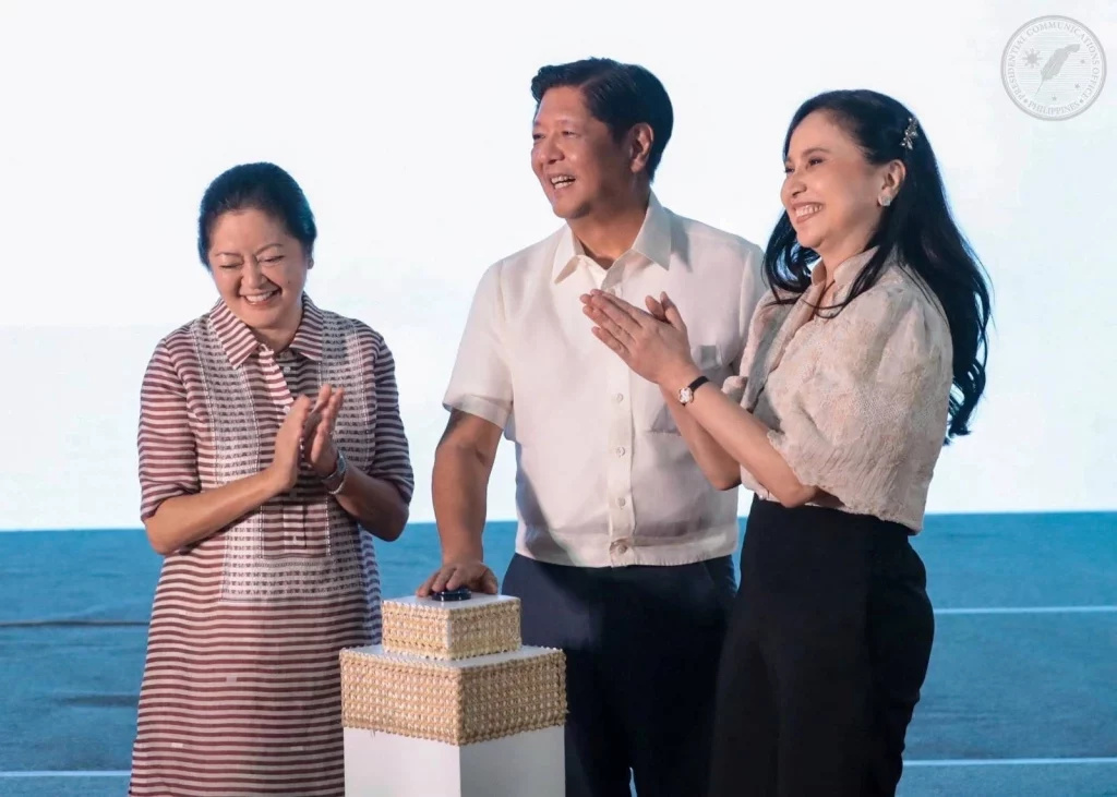 President Ferdinand Marcos Jr. (center), First Lady Liza Araneta Marcos (left), and DTI Secretary Cristina Roque (right) smile and applaud during the opening ceremony of the 2025 DTI Bagong Pilipinas National Food Fair. The President has his hand on decorative boxes placed on a podium.